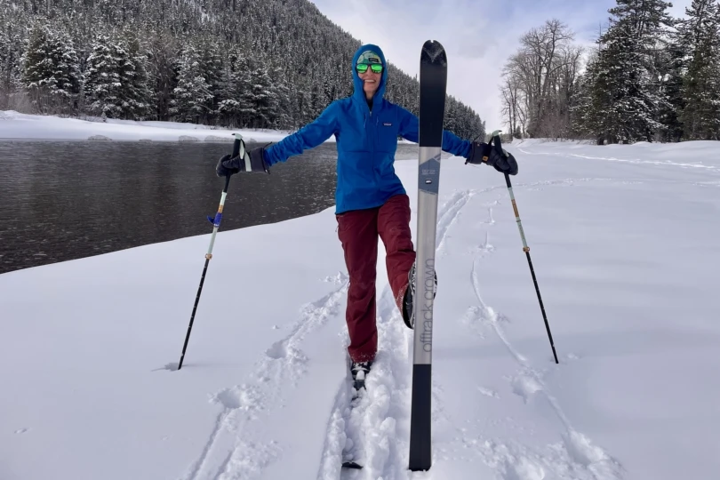A skier in a blue Patagonia R1 Thermal Hoody and red pants stands on snowy terrain by a river, holding ski poles and balancing one ski upright in front