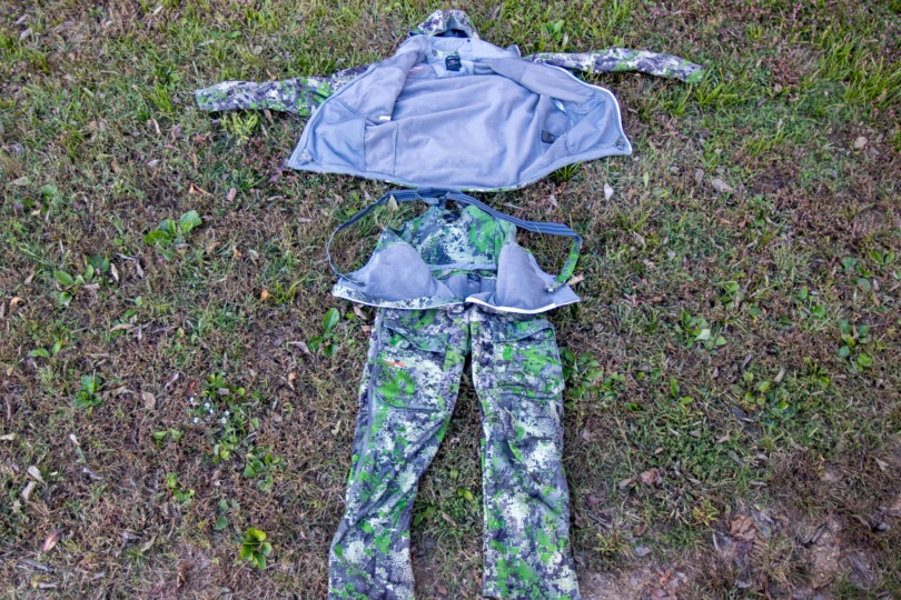 A hunting jacket and bib layed out on the grass.