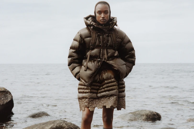 Person stands on rock in ocean wearing brown puffy clothing