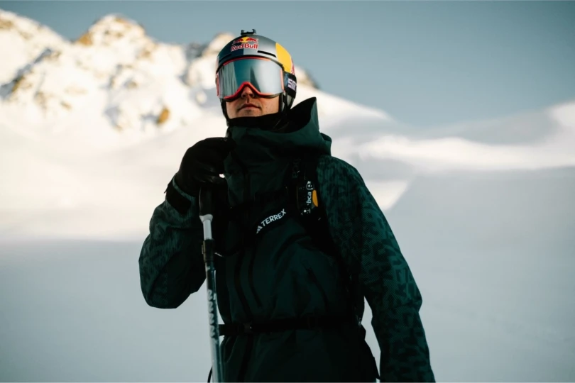 Skier stands wearing jacket and goggles on snowy mountain. 
