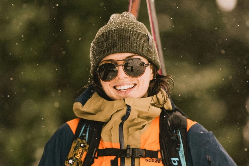 Person carrying skis in snow with beanie and sunglasses
