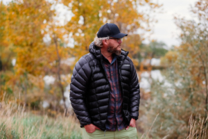 A man stands outdoors among autumn trees wearing a black puffer jacket and cap