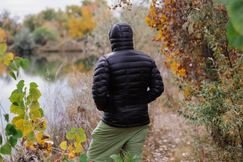 A man wearing a black hooded puffer jacket looks over a calm lake surrounded by fall colors
