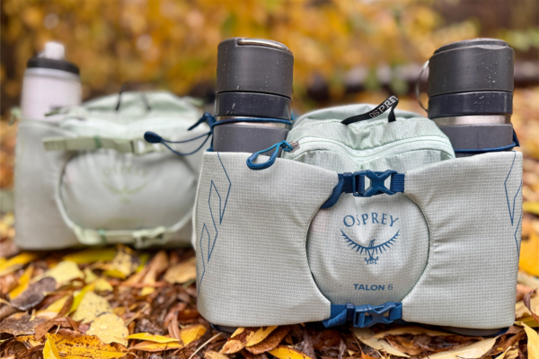 Two Osprey waistpacks side by side on fallen autumn leaves, each holding water bottles