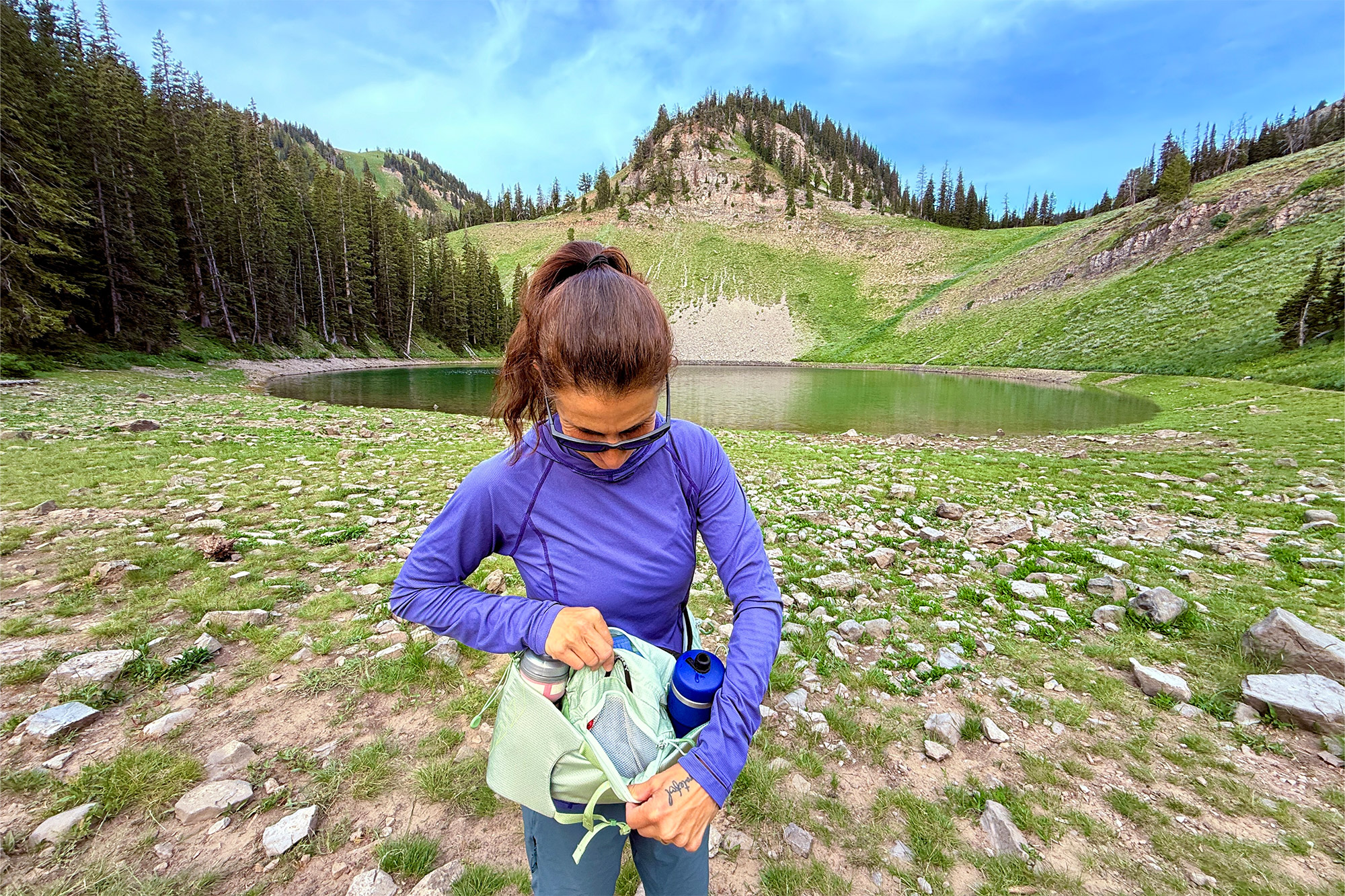 A woman adjusts items inside the waistpack while standing on rocky terrain by a calm alpine lake