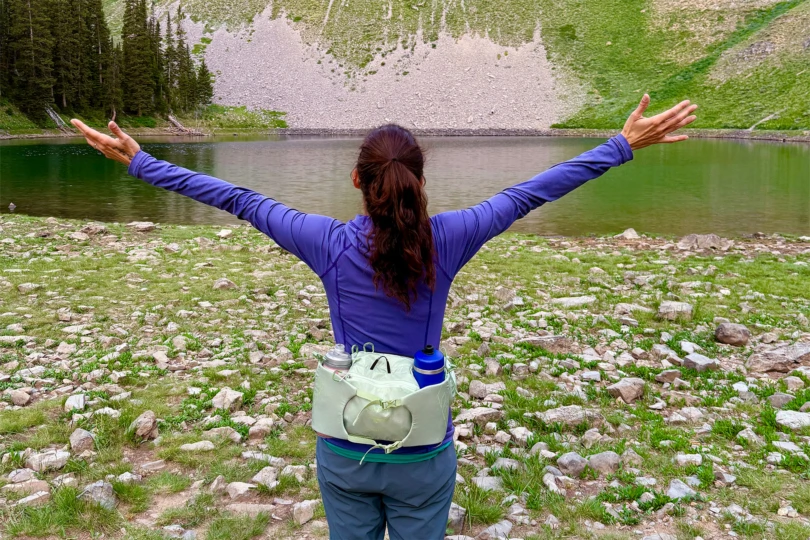 A hiker standing by a mountain lake with arms raised, wearing the Osprey Tempest & Talon 6 Waistpack