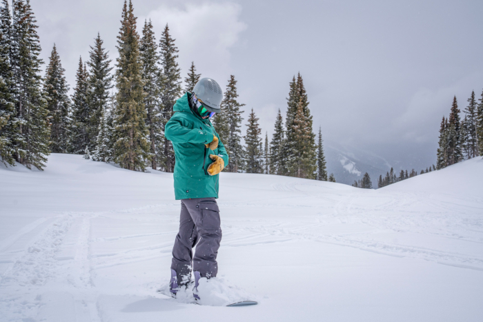 A snowboarder stands in deep snow adjusting a chest pocket on the 686 3-in-1 Rodeo Jacket