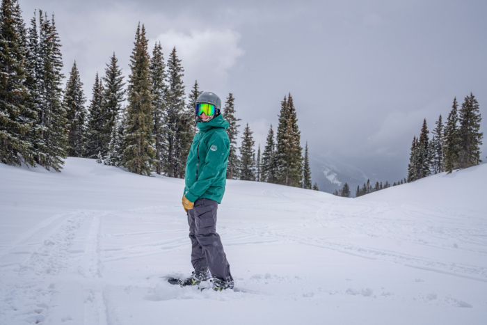 A rider stands on a snowy slope wearing the rodeo jacket with tall pine trees behind them