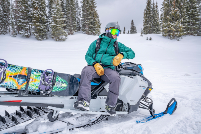 A snowboarder sits on a snowmobile wearing the 686 snowboard jacket, holding bright mittens