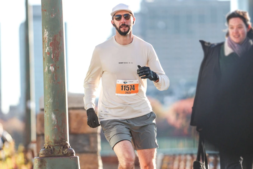 A man wearing sunglasses, gloves, and a white cap runs outdoors in cool weather with race bib number 11574 visible on his shirt
