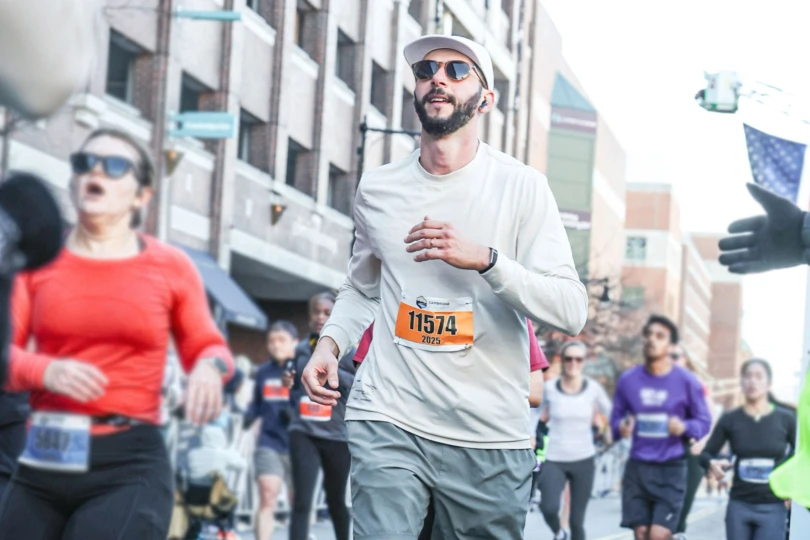 A man wearing sunglasses, a white running cap, and a long-sleeve shirt with bib number 11574 runs through a city street surrounded by other runners