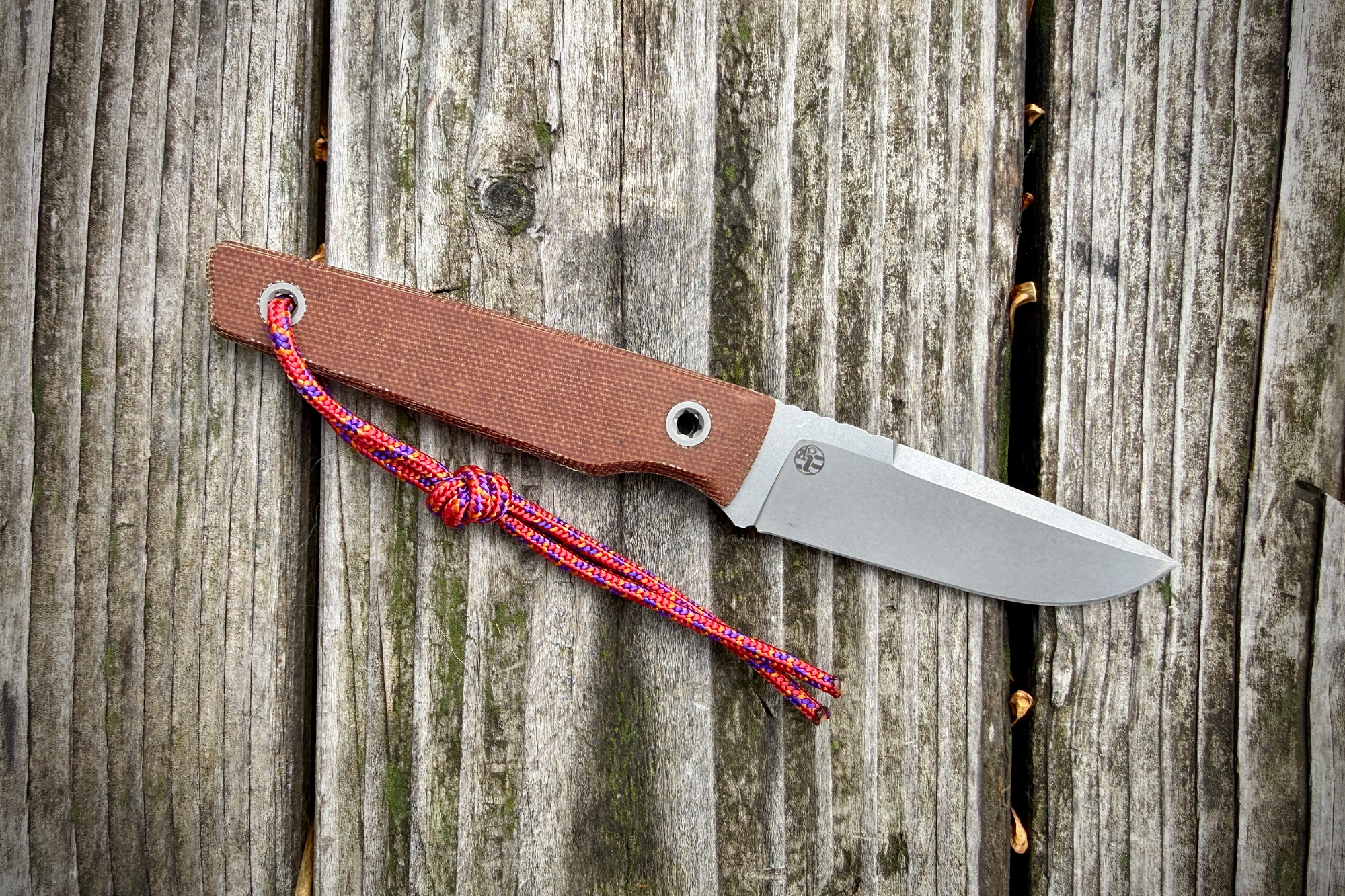 A small fixed blade knife lies on weathered wooden boards with a brown handle and bright red paracord