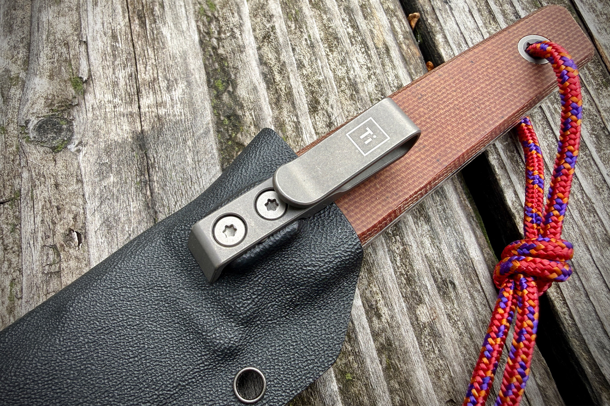 A close-up shows the metal pocket clip attached to the black sheath and the woven paracord hanging from the handle