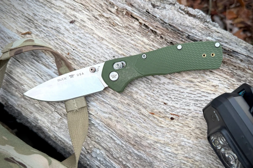 A Buck Range Elite knife with an olive handle resting on a log, blade open and catching the outdoor light