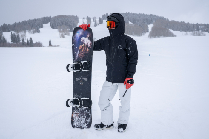 A snowboarder stands in the snow wearing the Burton [ak] Cyclic Jacket while holding a board upright on a snowy slope