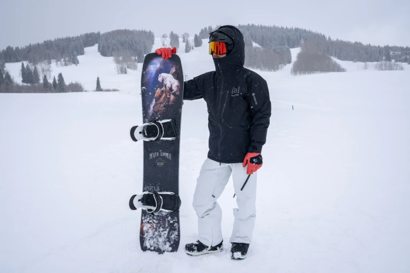 A snowboarder stands in the snow wearing the Burton [ak] Cyclic Jacket while holding a board upright on a snowy slope