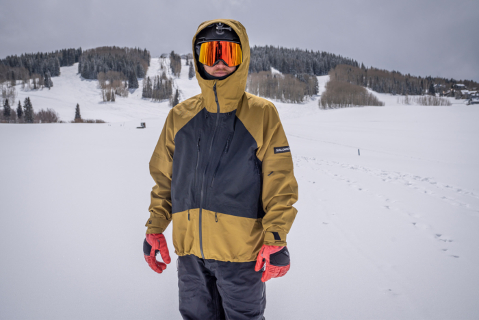 A rider stands in fresh snow wearing a salomon jacket with red gloves in front of a snowy mountain
