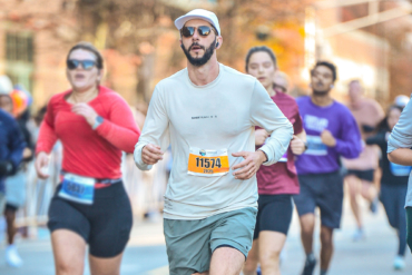 A man wearing sunglasses, a white running cap, and a light long-sleeve shirt with bib number 11574 runs among a group of racers on a sunny day
