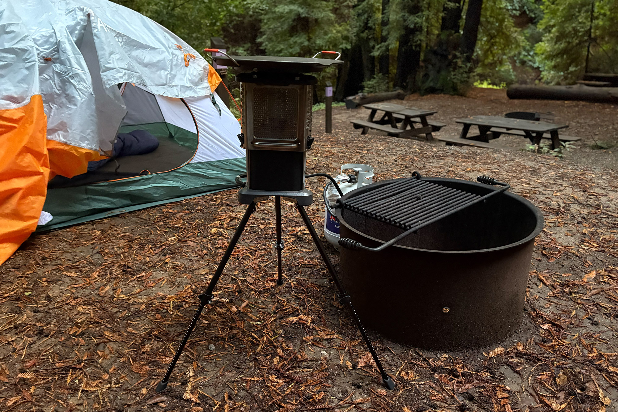 The Ignik Skullet Heater Stove stands on its tripod next to a tent and a metal fire ring at a forest campsite