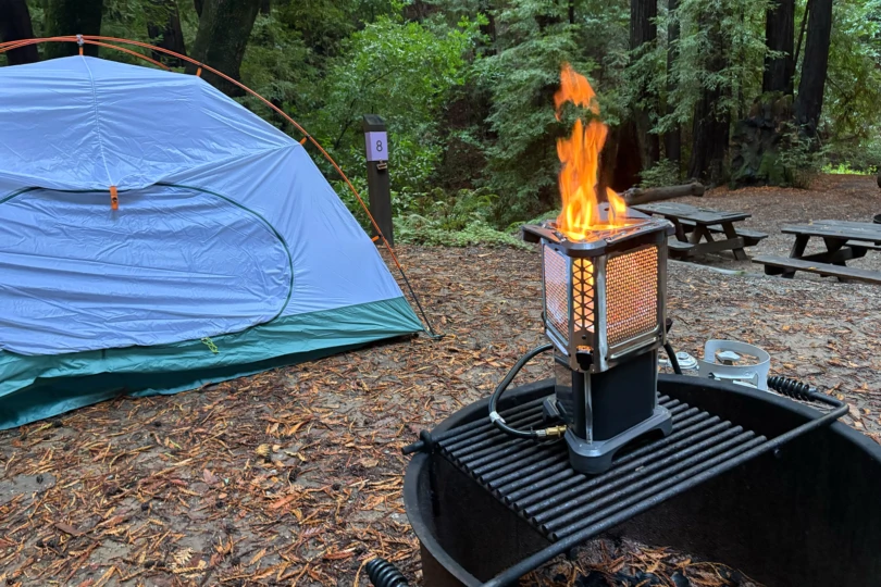 The heater stove burns with tall flames next to a pitched tent in a forest campsite