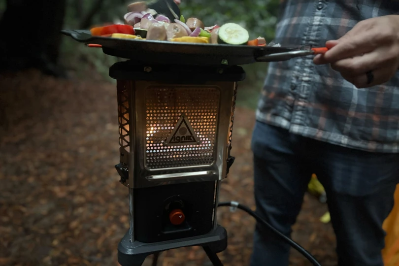The Ignik Skullet Heater Stove glows while a person cooks vegetables on a pan above it at the campsite