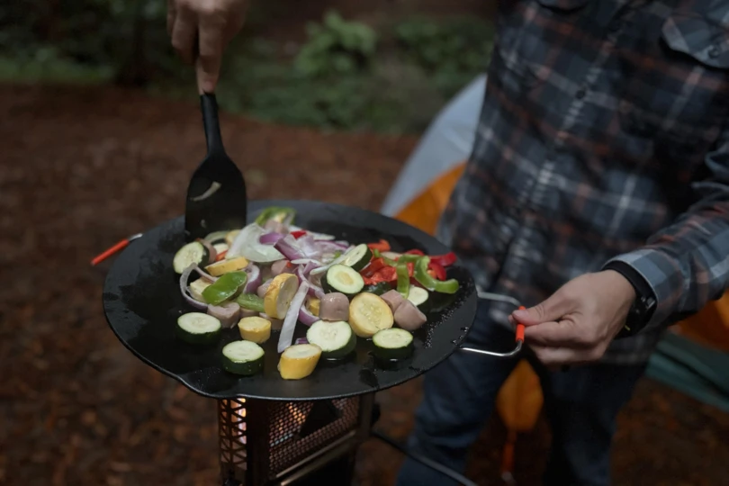 Mixed vegetables and sausage pieces cook on a flat pan over the heater stove while a person in a plaid shirt stirs them