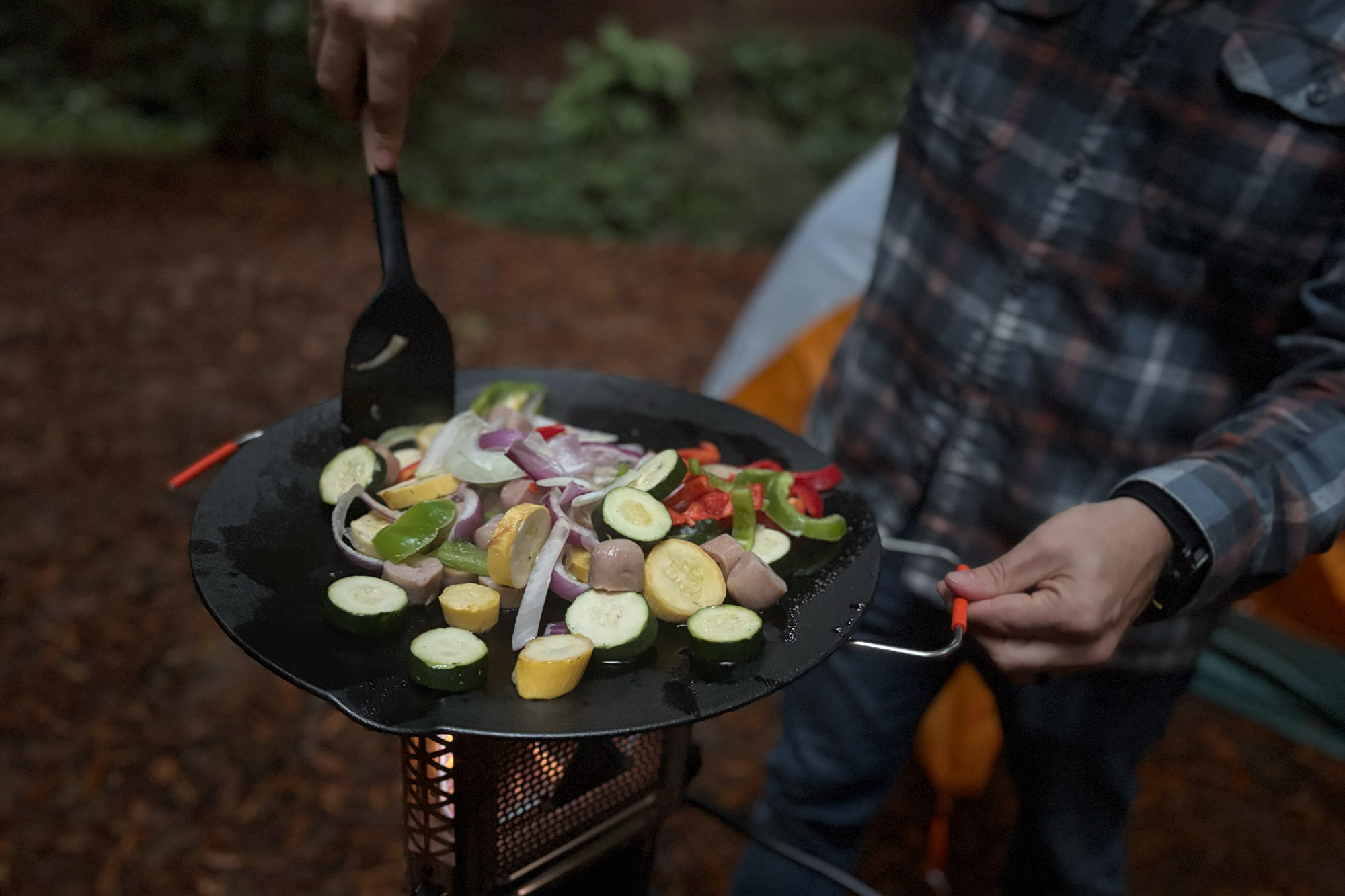Mixed vegetables and sausage pieces cook on a flat pan over the heater stove while a person in a plaid shirt stirs them