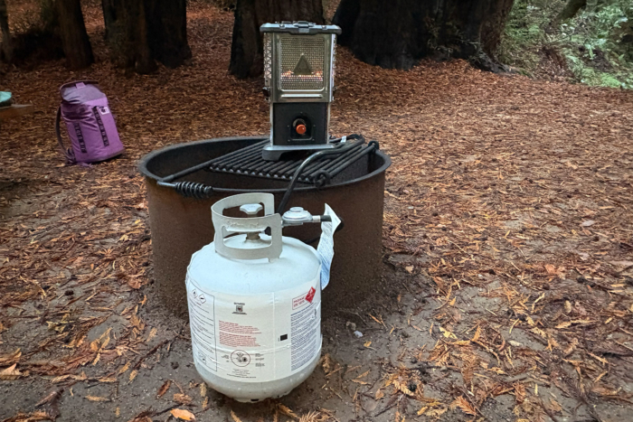 A propane tank sits beside the heater stove placed on top of a metal fire ring in a wooded campsite