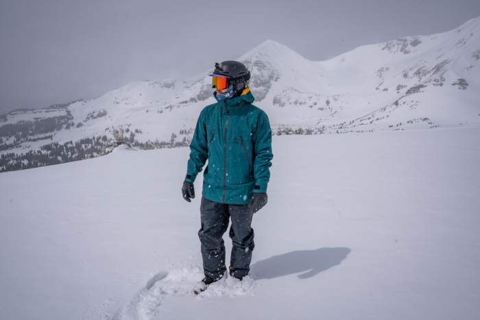 A snowboarder stands in fresh snow wearing the Jones Shralpinist jacket with mountains behind