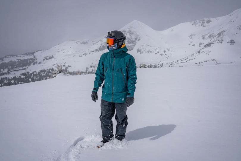 A snowboarder stands in fresh snow wearing the Jones Shralpinist jacket with mountains behind