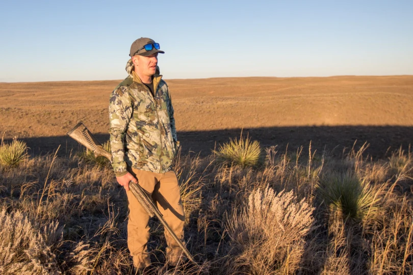 A hunter on top of a hill in the plains holding a shotgun.