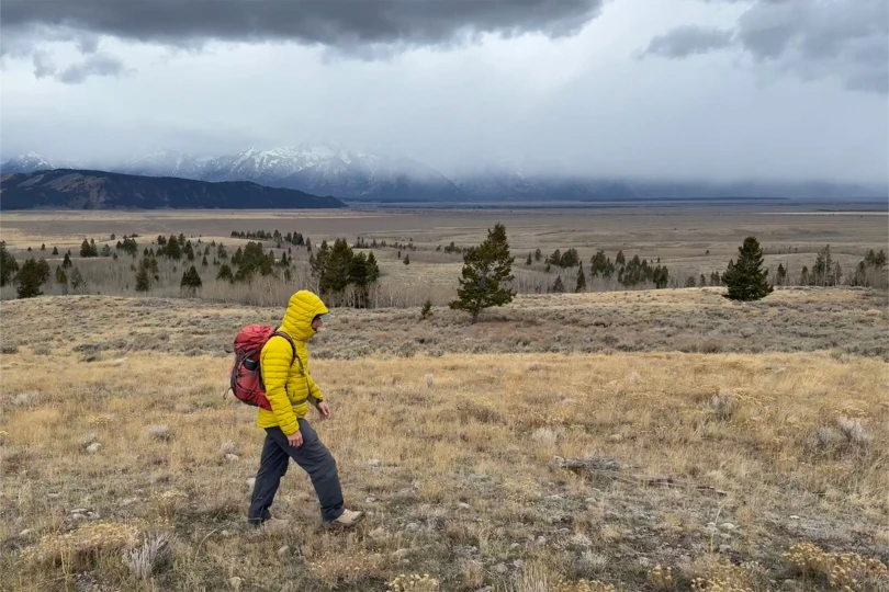 Hiker in a yellow Rab Microlight Windstopper and red backpack walking under dark clouds across open terrain