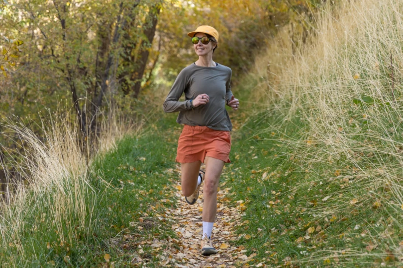 A runner moves uphill on a narrow forest trail wearing orange shorts and a long-sleeve top