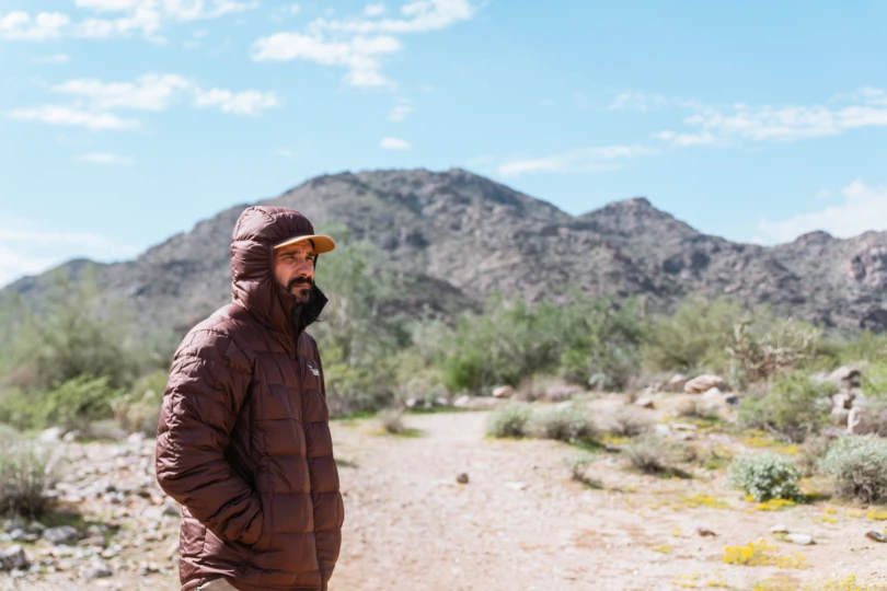 A man stands on a desert trail wearing the Sitka MicroDown Hooded Jacket with the hood up, with mountains and blue sky behind him
