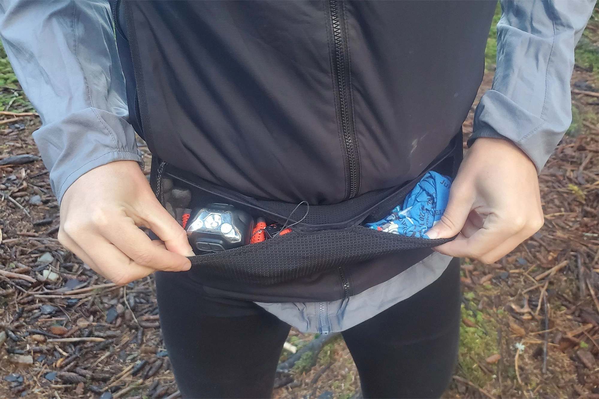 A person opening the running belt’s front pocket to show stored items including a headlamp and a blue cloth