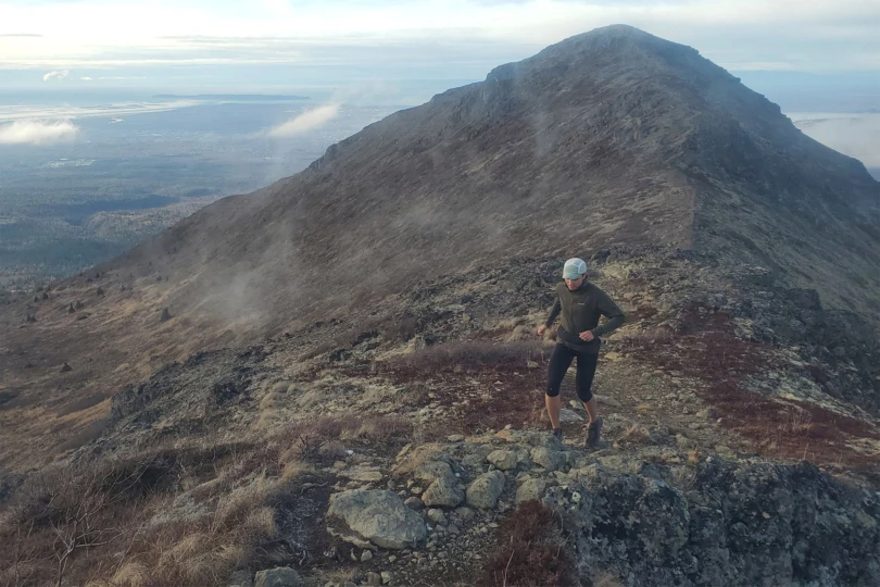 A runner moves along a rocky ridgeline high in the mountains, with a long, rugged slope stretching behind them and patches of fog drifting across the landscape. The running belt is worn around their waist