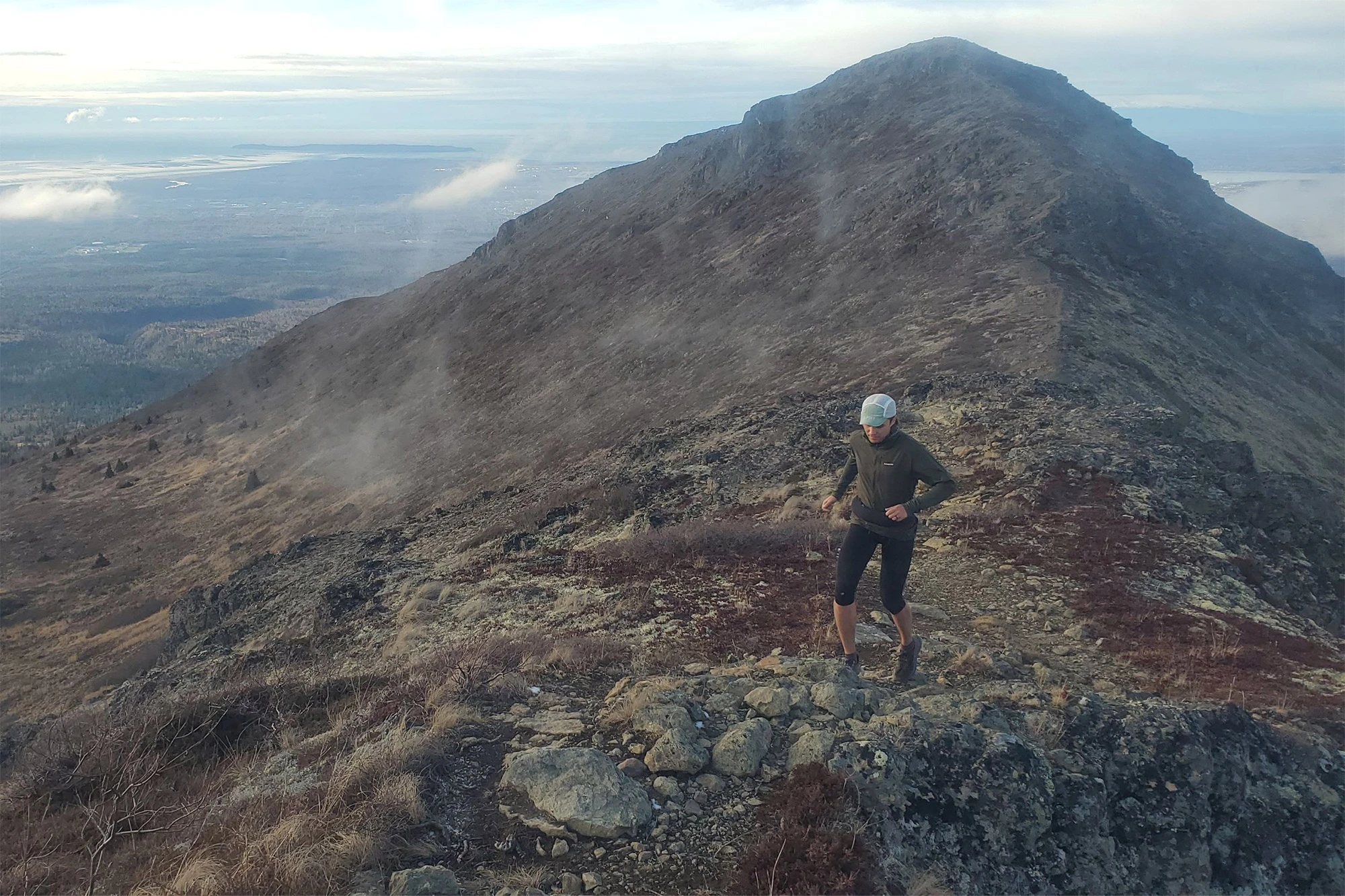 A runner moves along a rocky ridgeline high in the mountains, with a long, rugged slope stretching behind them and patches of fog drifting across the landscape. The running belt is worn around their waist