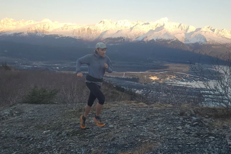 A trail runner mid-stride on rocky terrain, with The Raide LF 2L visible around their waist as snowy mountains rise in the background