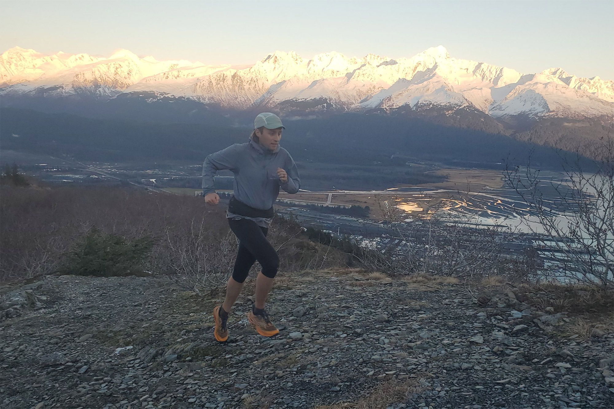 A trail runner mid-stride on rocky terrain, with The Raide LF 2L visible around their waist as snowy mountains rise in the background