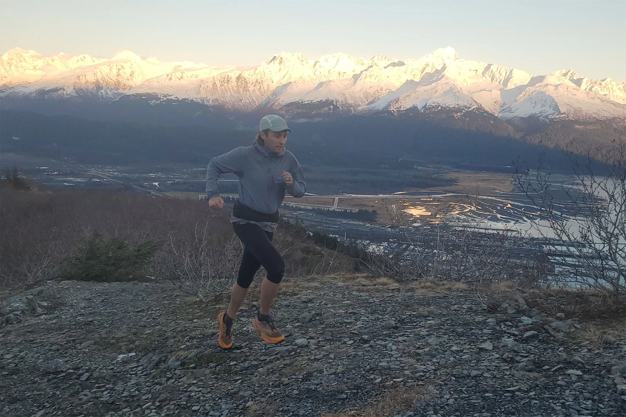 A trail runner mid-stride on rocky terrain, with The Raide LF 2L visible around their waist as snowy mountains rise in the background