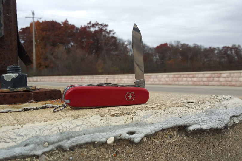 A red Victorinox Swiss Army Companion sits on a concrete ledge with its main blade open, with fall trees blurred in the background