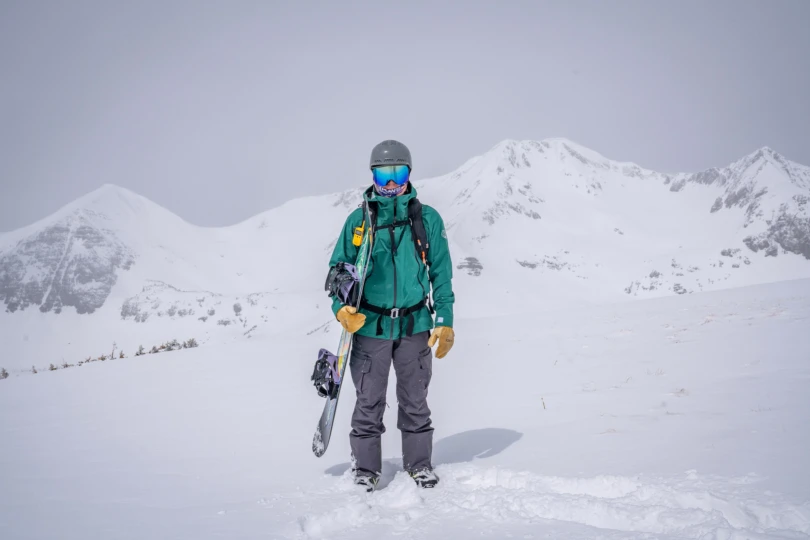 A snowboarder stands in an open snowy basin holding a board while wearing the 686 3-in-1 Rodeo Jacket