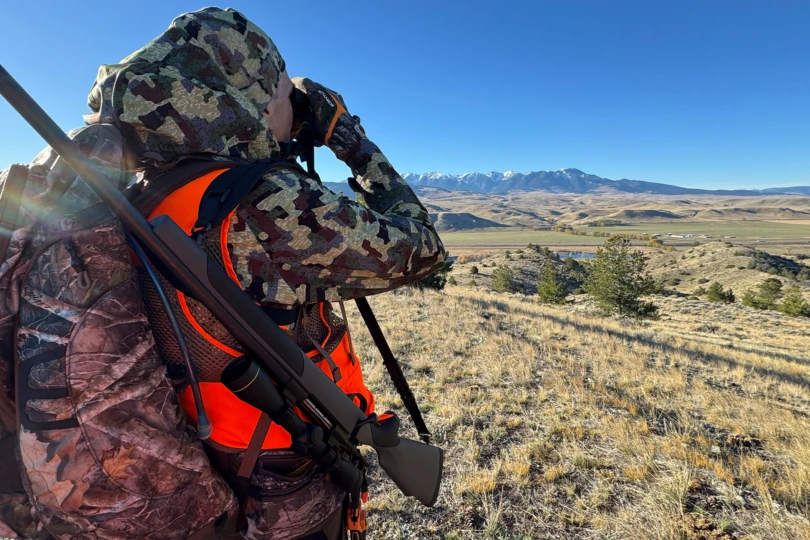 A camo soft shell jacket is worn while using binoculars on a grassy hillside