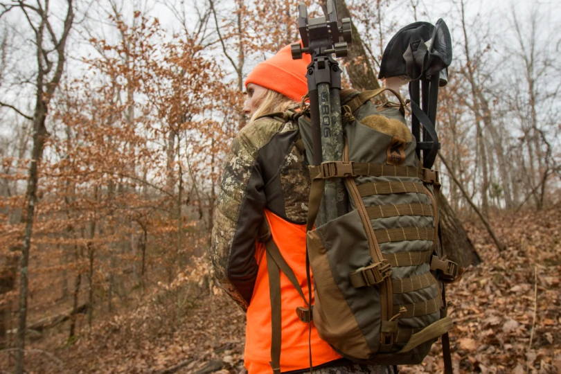 A female hunter carrying a pack with a seat and a shooting tripod attached to it.