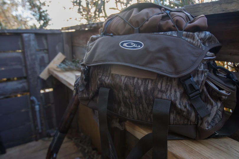 The Drake Waterfowl Large blind bag sitting on a shelf in a duck blind next to an over/under shotgun.