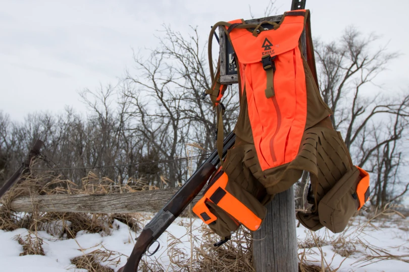 The Chief Upland Over/Under Vest hanging on a post next to an over under shotgun in front of a winter landscape.