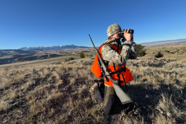 A hunter kneels while wearing an AllClima jacket and using binoculars on a ridge