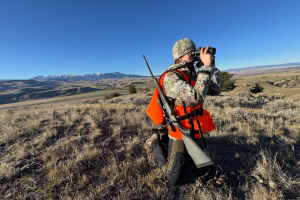 A hunter kneels while wearing an AllClima jacket and using binoculars on a ridge