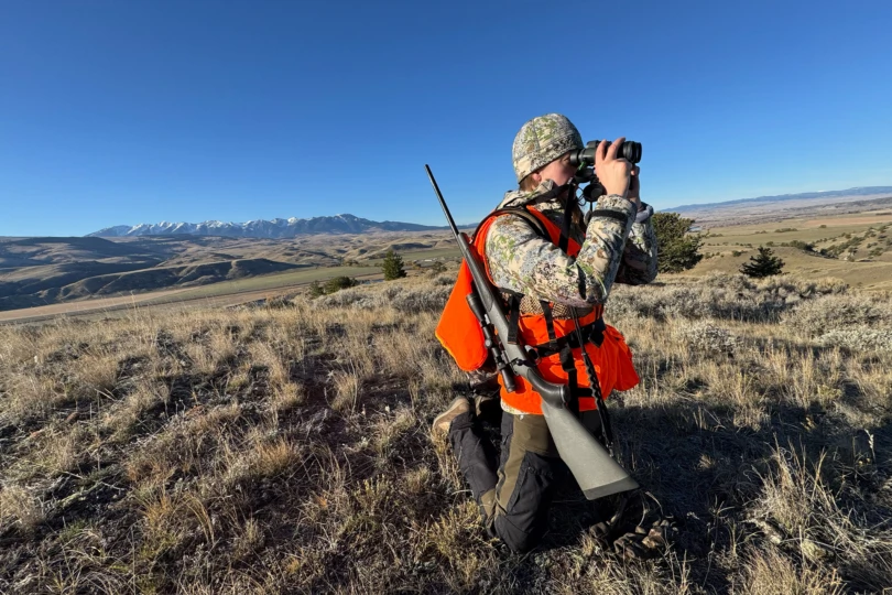 A hunter kneels while wearing an AllClima jacket and using binoculars on a ridge