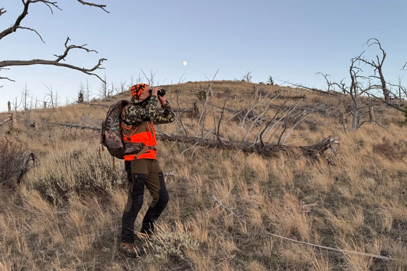 A hunter stands glassing a hillside with binoculars while wearing reinforced pants